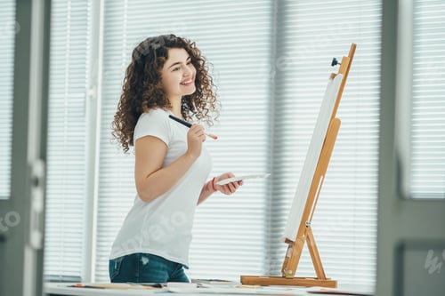 Preview: The happy curly woman painting a picture on the easel.