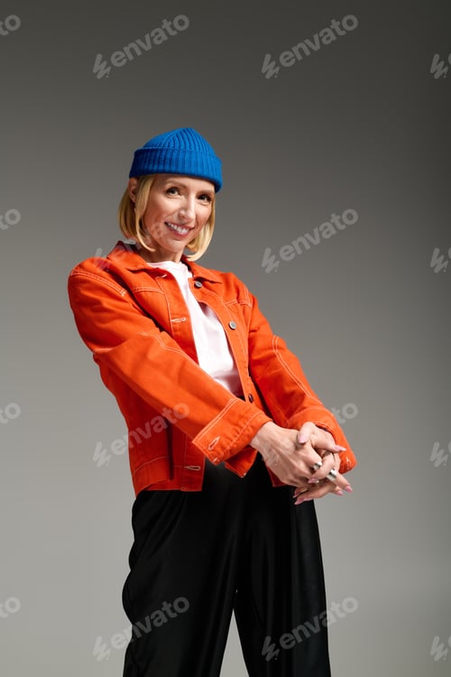 Preview: Stylish blonde woman in blue beanie and orange jacket showcases modern fashion in studio