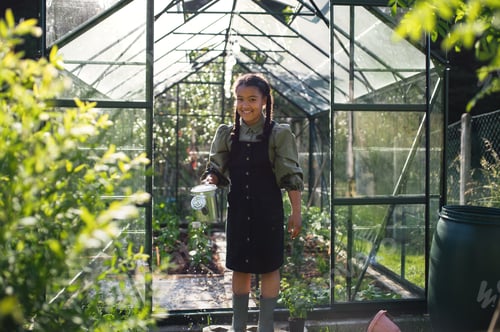 Preview: Happy small girl gardening in greenhouse outdoors in backyard, looking at camera