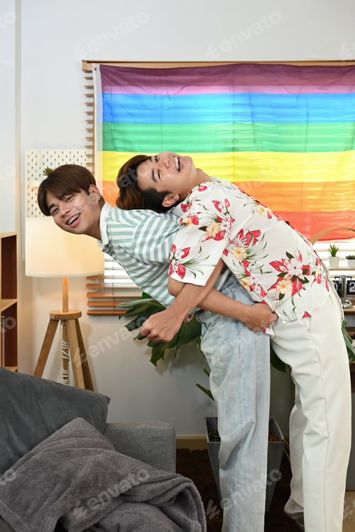 Preview: Cheerful gay male couple giving piggyback ride for each other laugh joyfully in living room