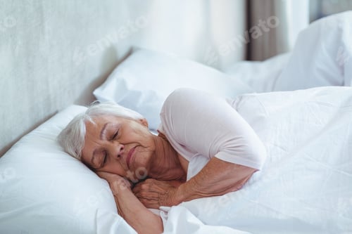 Preview: Senior woman lying on side in bed wearing sleep top and hugging white pillow against headboard