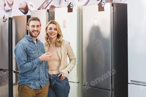 Preview: smiling girlfriend and boyfriend pointing with finger at fridge in home appliance store