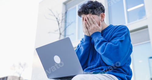 Vista previa: Estudiante estresado que se cubre la cara con las manos mientras trabaja en una computadora portátil fuera de la universidad
