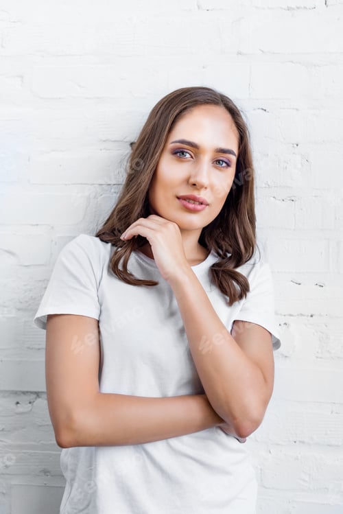 Preview: beautiful young woman in white t-shirt standing near white brick wall and looking at camera