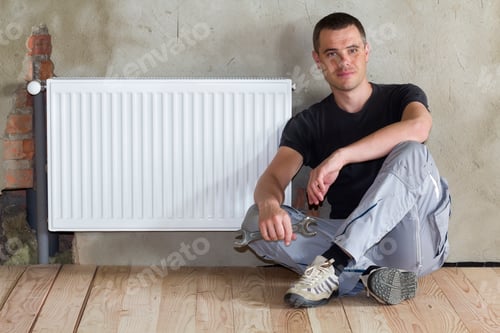 Preview: Young handsome plumber sitting on floor with wrench in hand near successfully installed heating