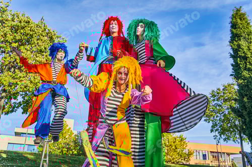 Preview: Cheerful harlequins on stilts and unicycle in park during masquerade