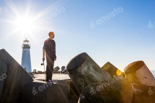 Preview: Young man holding skateboard, standing on breakwater near lighthouse, looking towards ocean