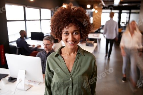 Preview: Millennial black female creative standing in a busy casual office, smiling to camera