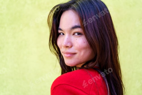 Preview: Young asian woman in vibrant red sweater turning head, smiling brightly against green backdrop