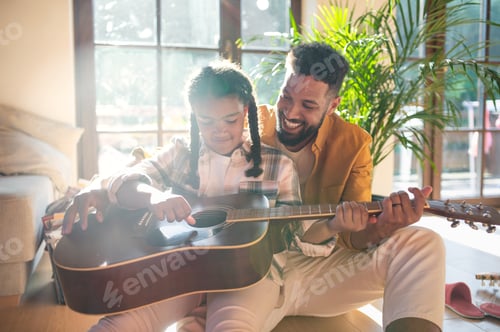Visualização: Vista frontal do pai feliz com a filha pequena dentro de casa, tocando violão