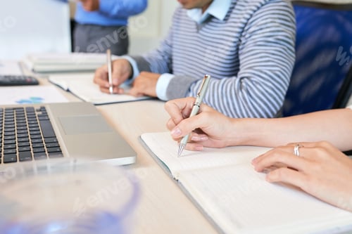 Preview: Crop coworkers writing in notepads at table
