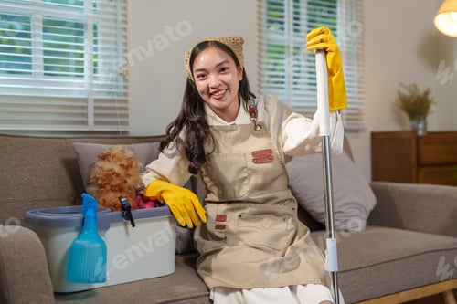 Preview: Young asian housekeeper smiling while taking a break on the sofa, holding a mop and with cleaning su