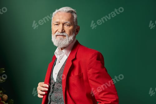 Preview: portrait of stylish man in red Santa suit looking at camera on green backdrop, winter concept