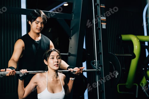 Preview: Asian woman guy a squat exercise legs heavy weights with a barbell with the assistance of a trainer