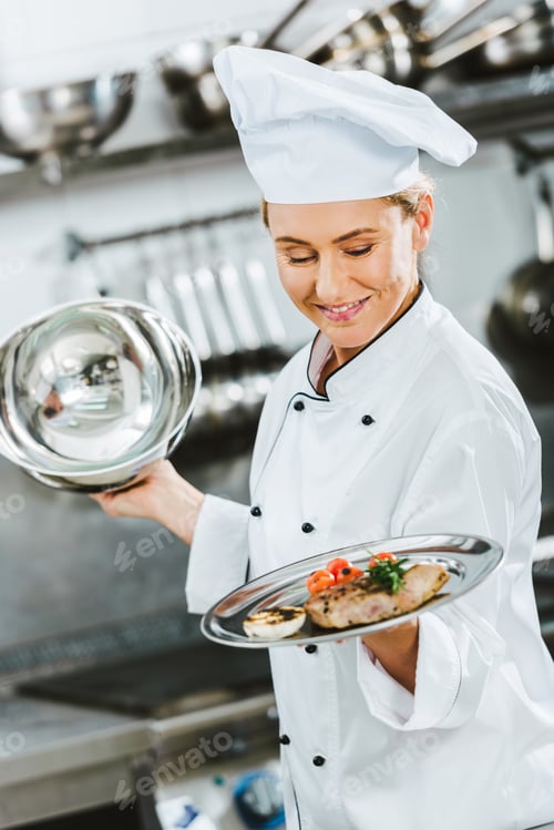Preview: selective focus of beautiful female chef in uniform holding serving tray with meat dish in