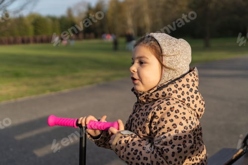 Preview: Caucasian preschool girl portrait with pink scooter in the park on sunny cold day in spring