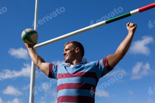 Preview: Smiling rugby player holding ball with arms raised against blue sky