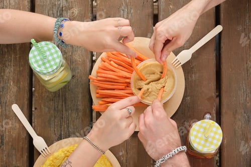 Preview: Group of unrecognizable happy friends taking hummus with carrot sticks in a park having picnic