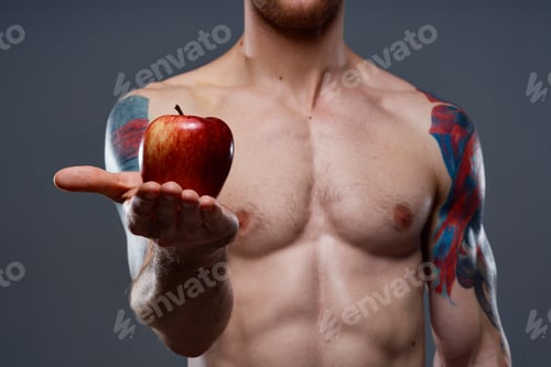 Preview: tattooed athlete holding red apple on gray background naked torso cropped view
