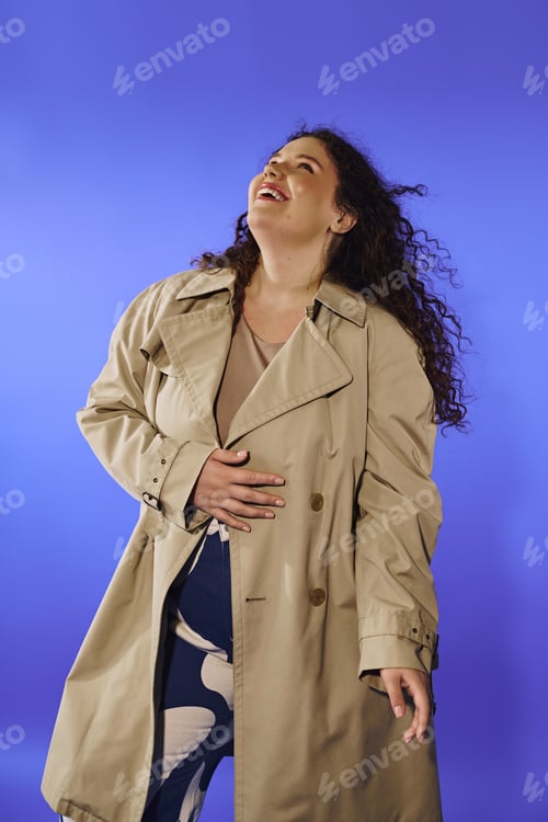 Preview: Joyful plus size woman in a stylish coat laughing against a vibrant blue backdrop