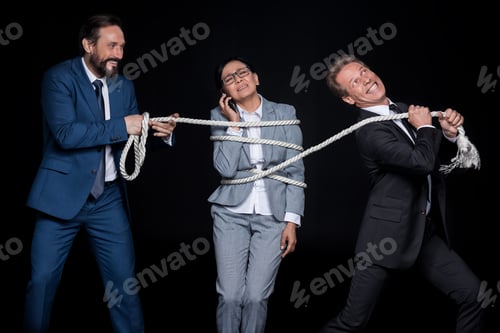 Preview: smiling businessmen with rope tying mature scared asian businesswoman talking on smartphone