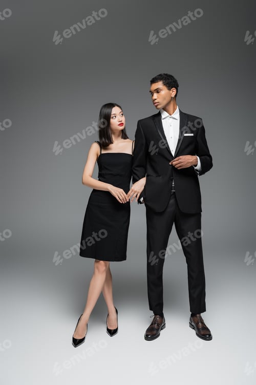 Preview: full length of interracial couple in black elegant attire looking at each other on grey background