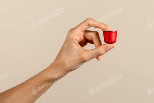 Preview: Unrecognizable woman hand showing coffee capsule against beige backdrop