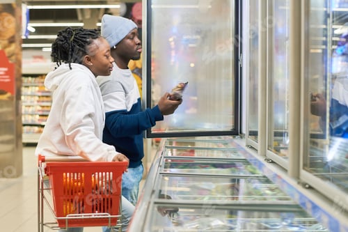 Preview: Young couple with shopping cart choosing frozen food products in supermarket