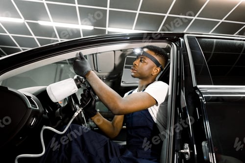 Preview: Professional chemical cleaning of interior of luxury car by air gun. Male African worker of auro
