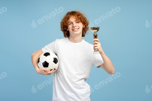 Preview: Happy young man with braces, wearing sport clothing, raising a trophy and holding soccer ball