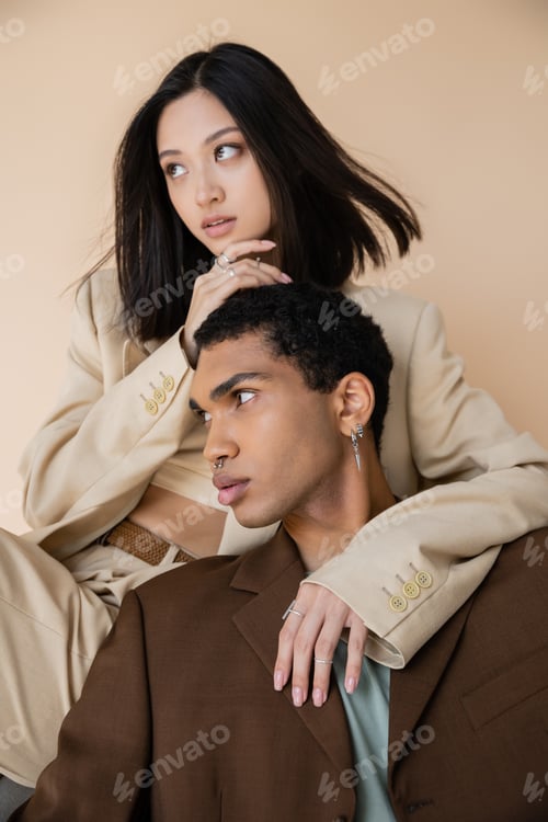 Preview: fashionable interracial couple in blazers looking away isolated on beige