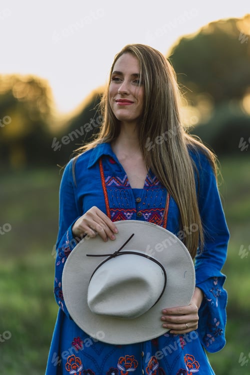 Preview: Pretty Caucasian young woman in a blue dress and a hat standing in a sun-kissed meadow