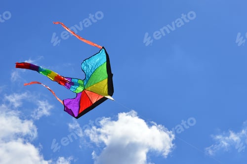 Preview: Rainbow colored kite flying on a clear summer day against a bright blue sky with puffy clouds