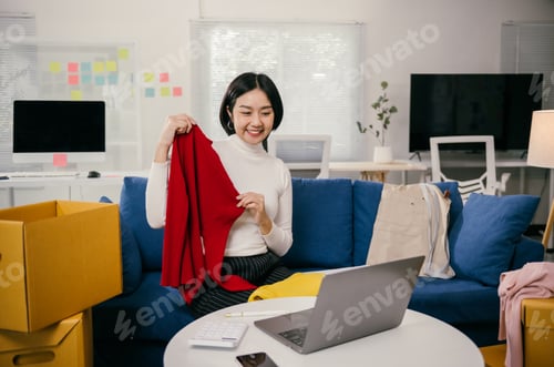 Preview: Asian female entrepreneur showing clothes in a live stream using laptop at home