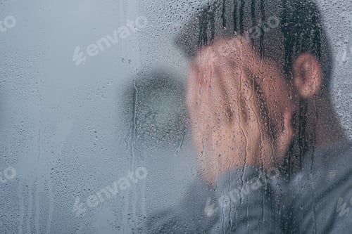 Preview: selective focus of raindrops on window with man covering face and crying on background