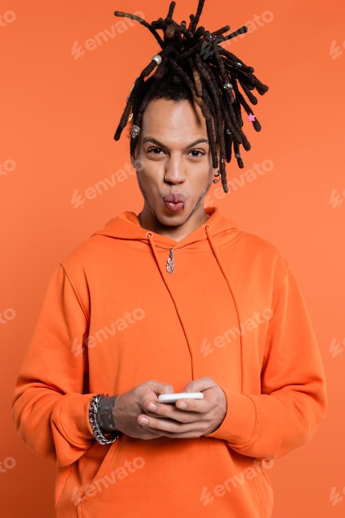 Preview: tattooed multiracial man with dreadlocks pouting lips and holding cellphone isolated on coral