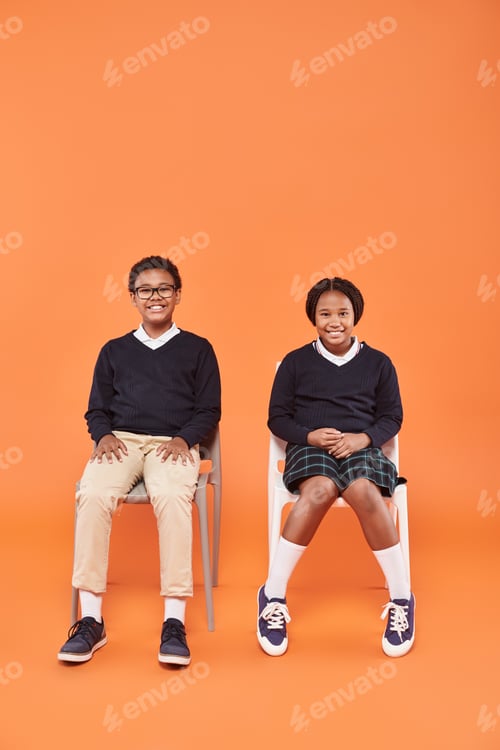 Preview: happy african american schoolkids in uniform smiling and sitting on chairs on orange backdrop