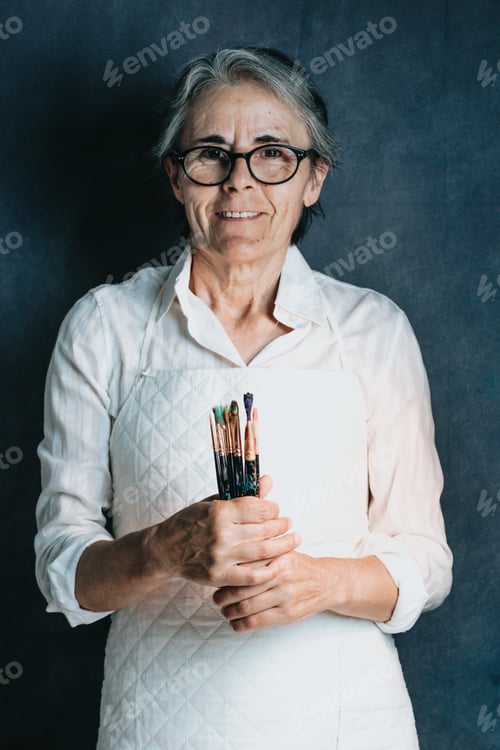 Preview: Smiling older woman, proud artist, with grey hair and glasses and many paintbrushes, artistic image