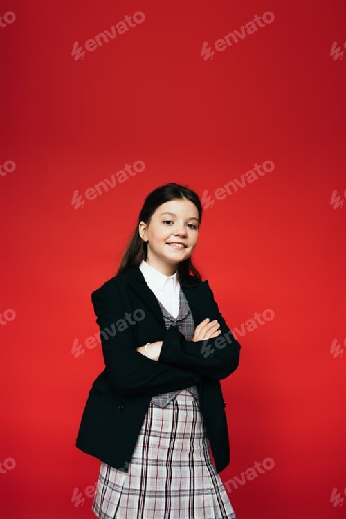 Preview: happy brunette child in black blazer and plaid skirt standing with crossed arms isolated on red