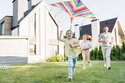 Preview: Happy daughter looking at camera while flying kite with parents running on background near house