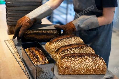 Preview: Close up of person wearing oven gloves placing freshly baked seeded loaves of bread on a tray in an