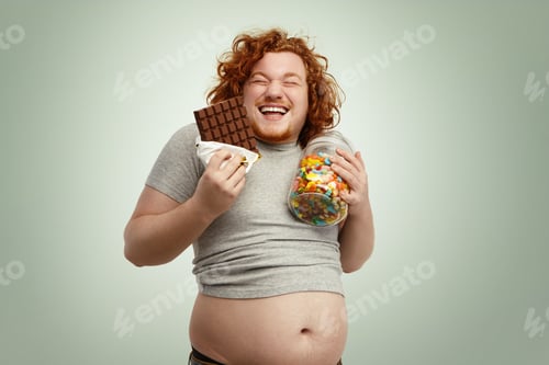 Preview: Indoor shot of fat redhead male holding glass of sweets in one hand and bar of chocolate in other, l