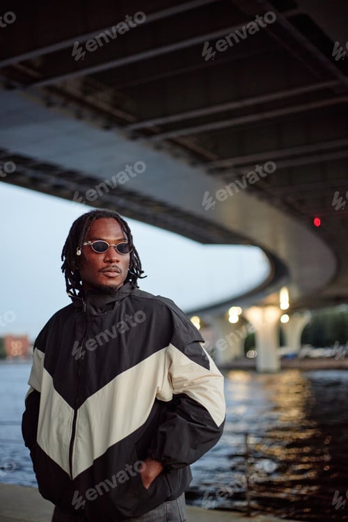 Preview: Young Adult Black Man Standing under Bridge Wearing Sunglasses at Dusk