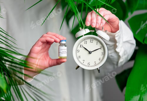 Preview: female hand holding alarm clock and vaccine in a bottle on background with palm leaves