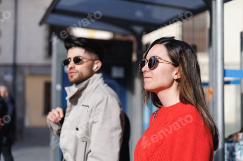 Preview: Woman and male friend waiting for the bus at the bus stop. The focus is on the woman.