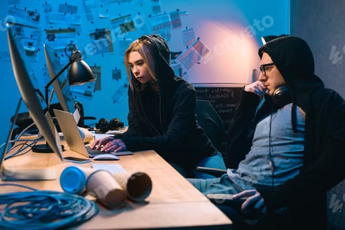 Preview: couple of young hackers using laptop at workplace in dark room