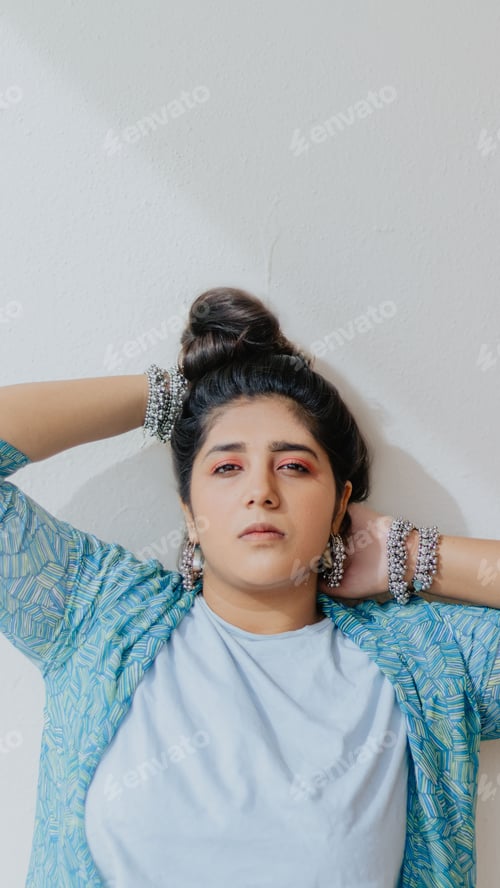 Preview: woman wearing blue sweater and standing against wall with hands on head