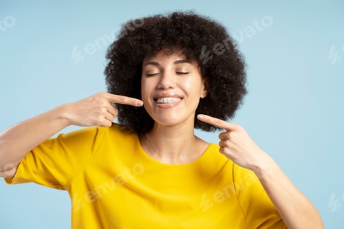 Preview: Smiling African American woman pointing invisible braces smiling with closed eyes on blue background