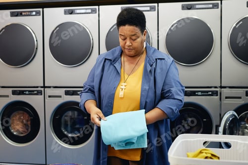Preview: Senior Black Woman Folding Clean Clothes after Washing in Public Laundry