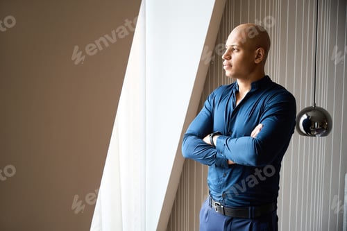 Preview: Thoughtful male entrepreneur with crossed arms looking in window in hotel room